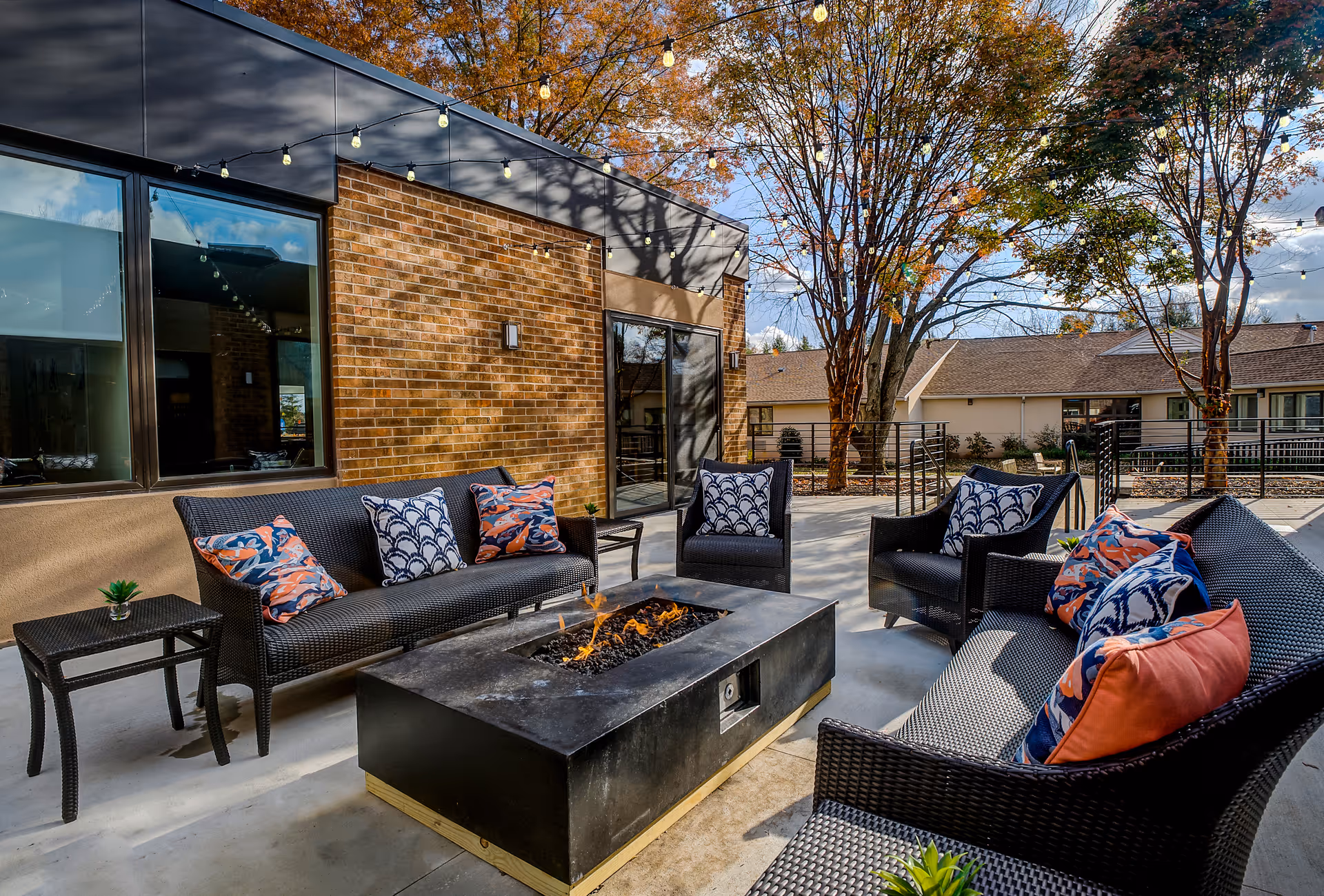 Outdoor patio area with black wicker seating including two sofas and two armchairs arranged around a rectangular fire pit table. The seating has colorful patterned cushions in blue, white, and orange. The patio is adjacent to a brick building with large windows and string lights overhead. Trees with autumn leaves are visible in the background.
