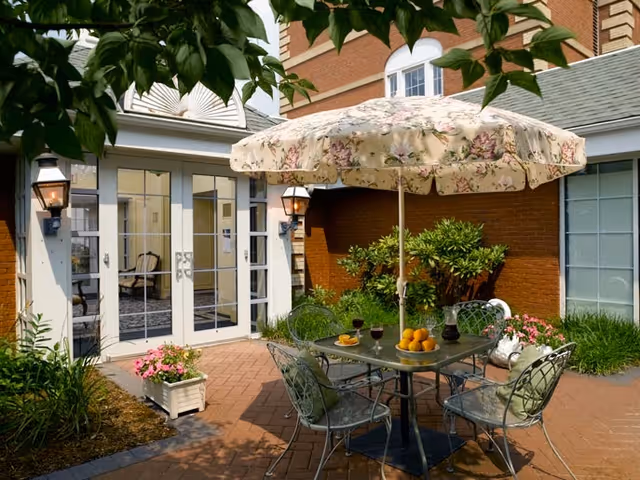 Sunny brick courtyard with a round metal table, four chairs and a floral umbrella outside glass double doors of the building.