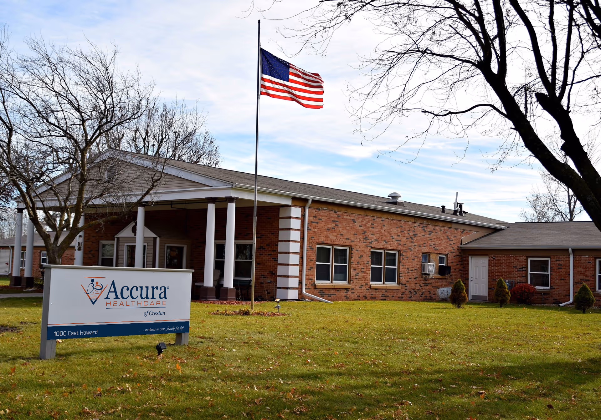 Single-story brick senior care building with an American flag on a pole, a lawn and a sign reading 'Accura HealthCare' in the foreground.