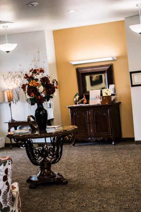 A cozy interior space with a round wooden table featuring intricate carvings, topped with a large vase of artificial flowers. In the background, there is a wooden cabinet with a mirror above it, some decorative items, and soft lighting. The room has patterned carpet and warm beige walls.