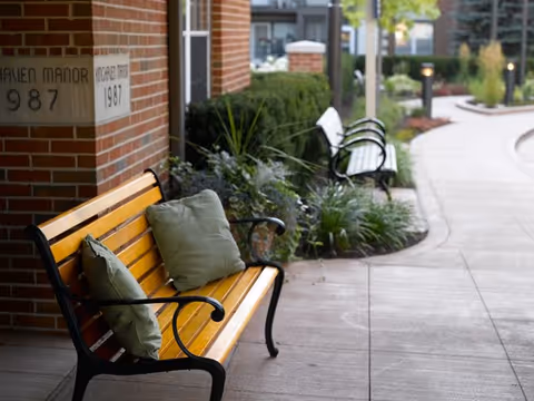 A wooden bench with green cushions placed along a paved walkway outside a brick building. The walkway curves and is lined with plants, bushes, and additional benches. The building has a sign that reads 'Kinghaven Manor 1987'.