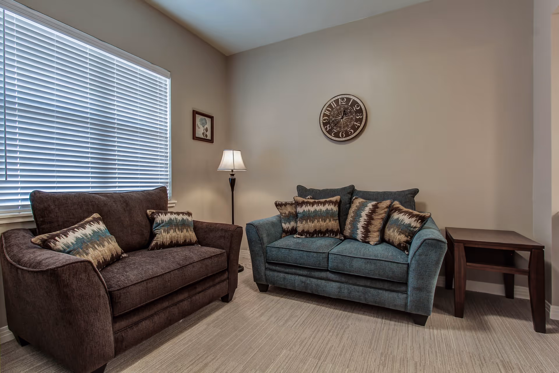 A cozy living room area with two upholstered loveseats, one brown and one teal, each adorned with patterned throw pillows. A floor lamp stands between the loveseats near a window with closed blinds. A round wall clock and a small framed picture hang on the beige walls. A wooden side table is placed next to the teal loveseat.