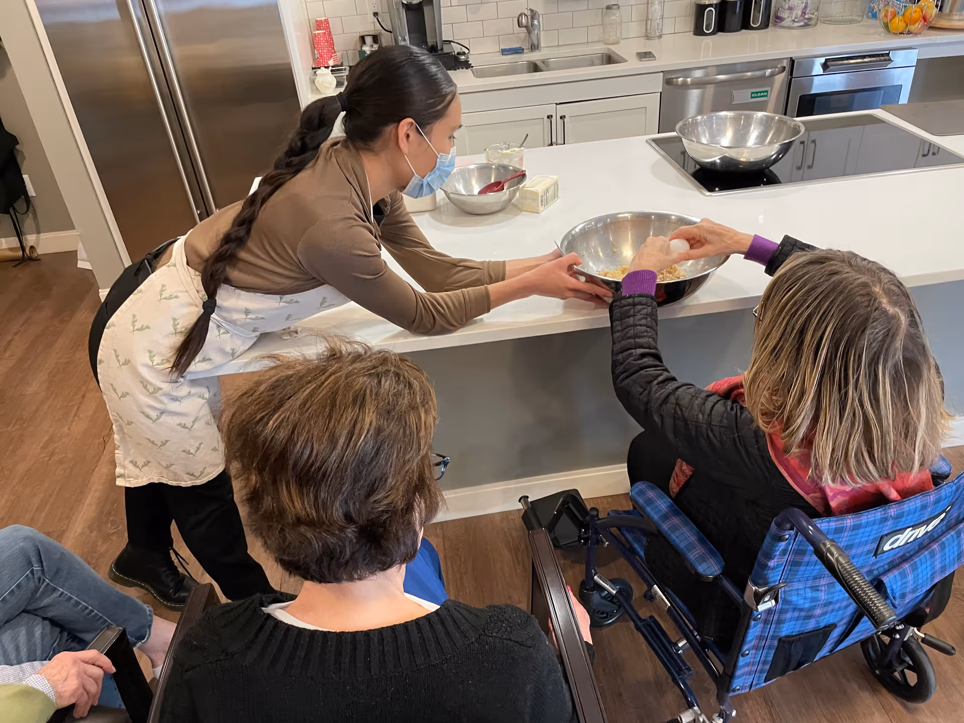A woman wearing a face mask and apron is assisting an elderly woman in a wheelchair with mixing ingredients in a large metal bowl on a kitchen counter. Two other elderly individuals are seated nearby, watching the activity in a bright kitchen with stainless steel appliances and wooden flooring.
