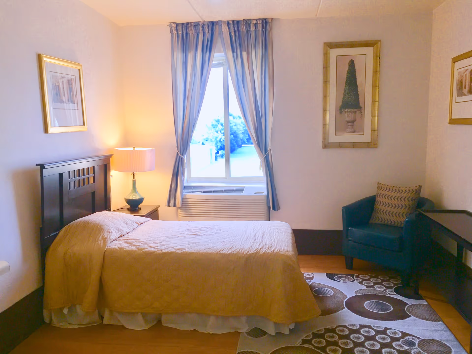 A cozy bedroom with a single bed covered in a beige quilt, a bedside table with a lamp, a window with blue and white curtains, a blue armchair with a patterned cushion, a dark wooden desk, and framed artwork on the walls.