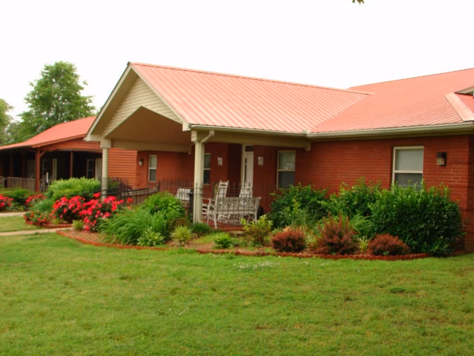 Exterior view of Oak Haven Assisted Care Living Facility showing a single-story brick building with a red metal roof, a covered porch with white rocking chairs, and well-maintained landscaping with green bushes and red flowers in front of the building.