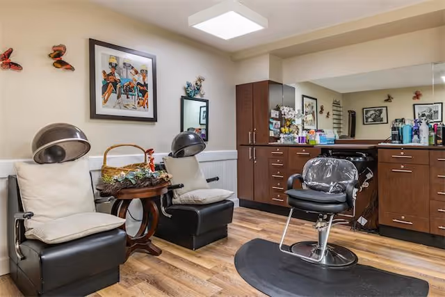 Interior view of a salon area in a senior living facility with two black hair drying chairs with white cushions, a small wooden table with a basket and decorative items, a black salon chair in front of a large mirror, and wooden cabinets with various hair care products on the countertop. The walls are decorated with framed artwork and butterfly decorations.