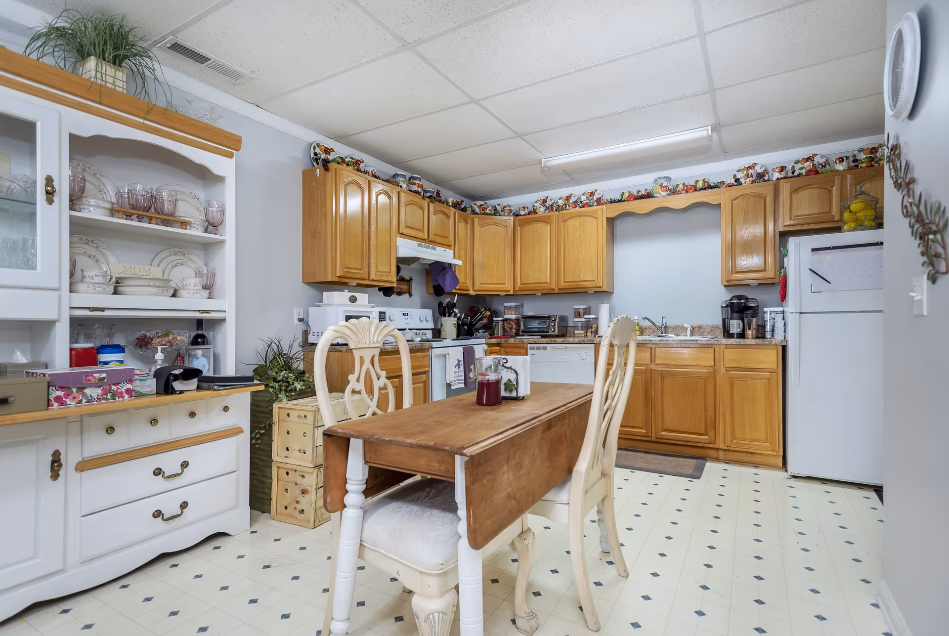 A cozy kitchen with wooden cabinets, a white stove, a white refrigerator, and a white dishwasher. There is a wooden table with two cream-colored chairs in the center. A white hutch with dishes and glassware is on the left side. The floor has a light-colored tile with a diamond pattern, and the ceiling has white tiles with fluorescent lighting. Decorative plates and containers are displayed on top of the cabinets.