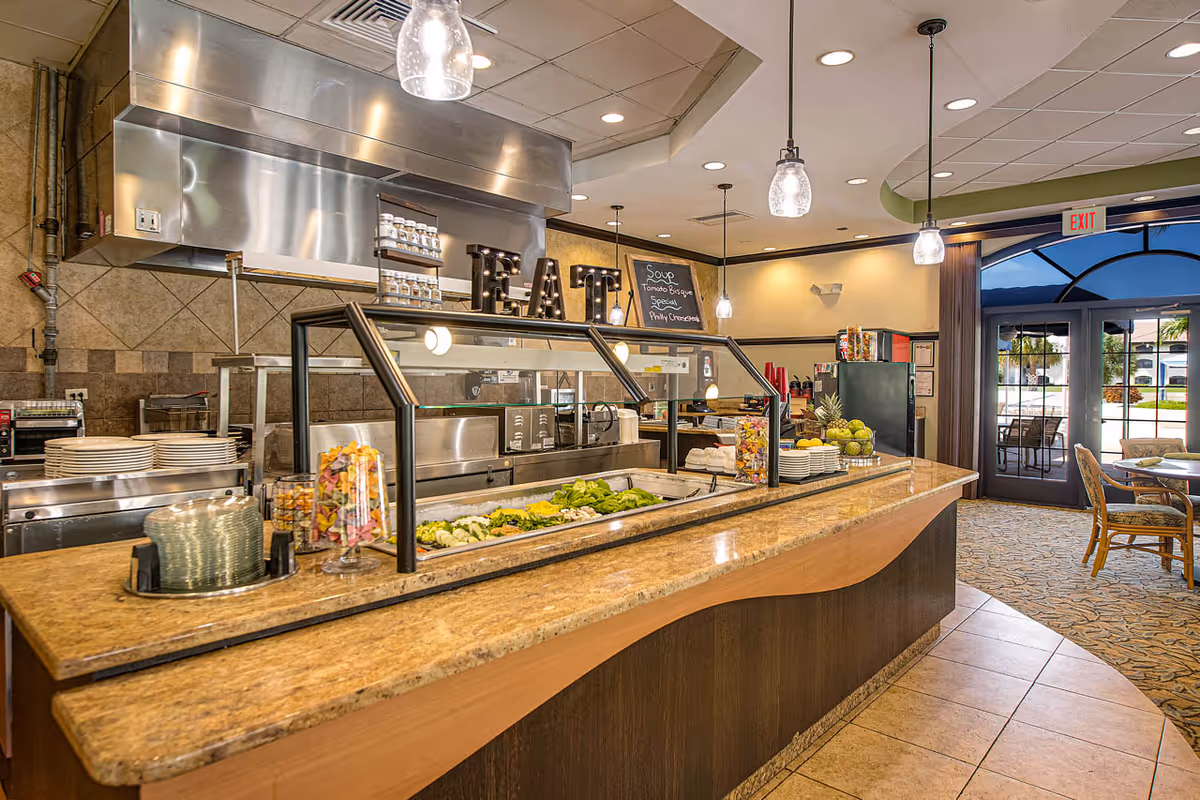 Interior view of a dining area with a buffet counter featuring a salad bar and plates. The counter has a granite top and a glass sneeze guard. Behind the counter, there is a kitchen area with stainless steel appliances and a sign that spells out 'EAT'. Pendant lights hang from the ceiling, and there is a chalkboard listing soups. In the background, there are tables and chairs near large glass doors leading outside.