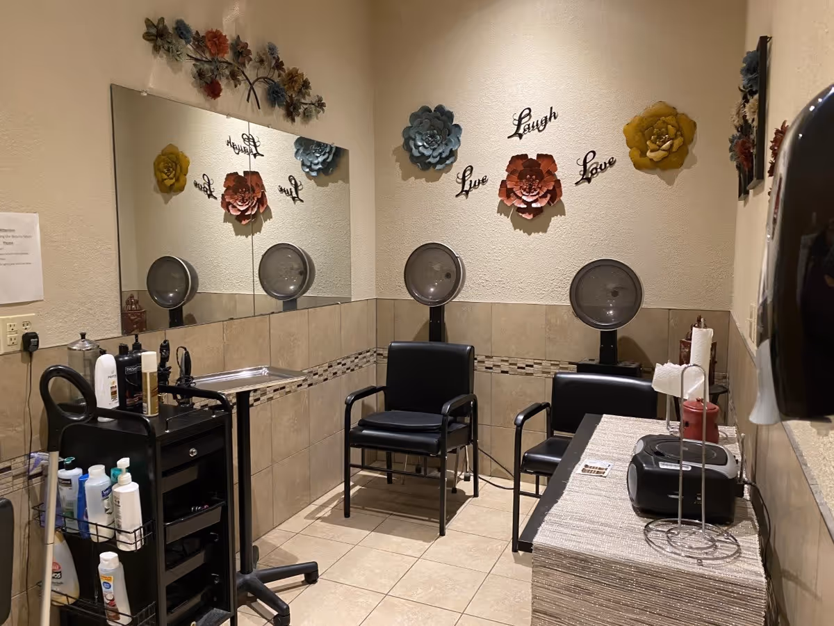 A small salon area with two black salon chairs facing wall-mounted hair dryers. The walls are decorated with large colorful flower wall art and the words 'Live Laugh Love'. There is a large mirror on one wall, a black rolling cart with hair care products, and a table with a paper towel holder and a small radio or speaker.