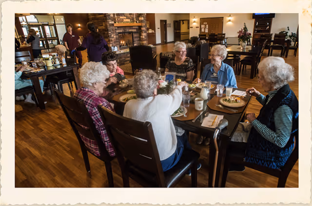 A group of elderly women sitting around a dining table enjoying a meal together in a warm, well-lit assisted living facility dining room. The room features wooden floors, a stone fireplace, and several other tables with chairs. Two staff members are visible in the background near the entrance.