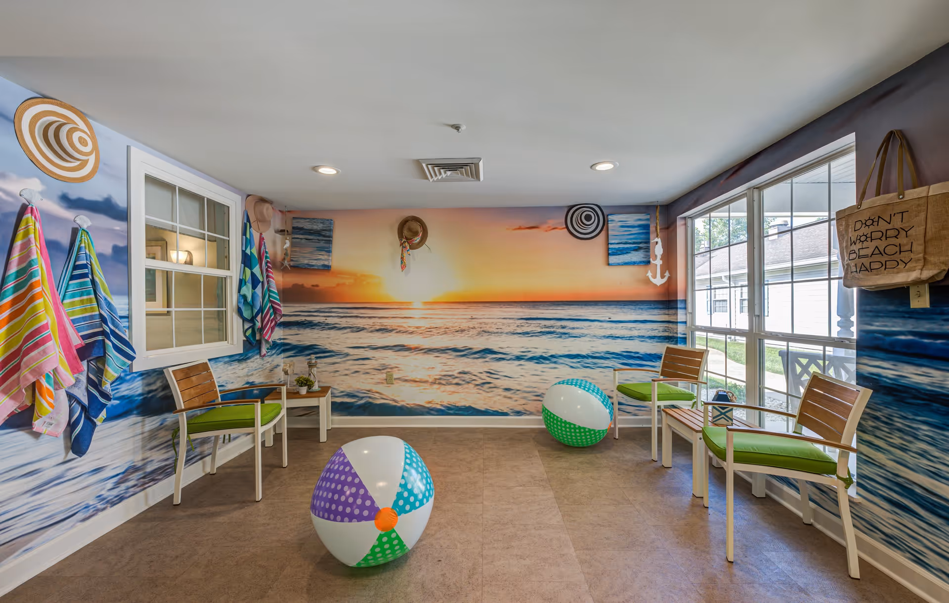 A bright room with walls decorated with a large mural of a beach sunset and ocean waves. The room contains four wooden chairs with green cushions, two colorful beach balls on the floor, and several striped beach towels hanging on hooks. There is a window on the left wall and large windows on the right side letting in natural light. A beige tote bag with the text 'DON'T WORRY BEACH HAPPY' hangs on the right wall.