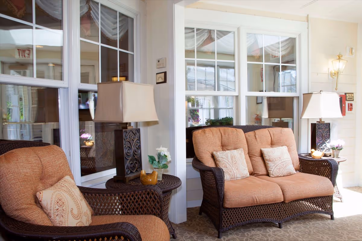 A cozy seating area with wicker furniture including a cushioned armchair and loveseat, both with beige cushions and patterned throw pillows. Two matching side tables hold decorative lamps and small flower arrangements. Large windows with white frames reflect the interior space, and a wall-mounted lantern-style light fixture is visible on the right.