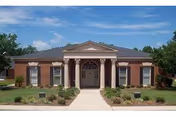 Front exterior view of a single-story brick building with a central entrance featuring four white columns and a triangular pediment. The building is surrounded by neatly trimmed bushes and small trees under a blue sky with some clouds.