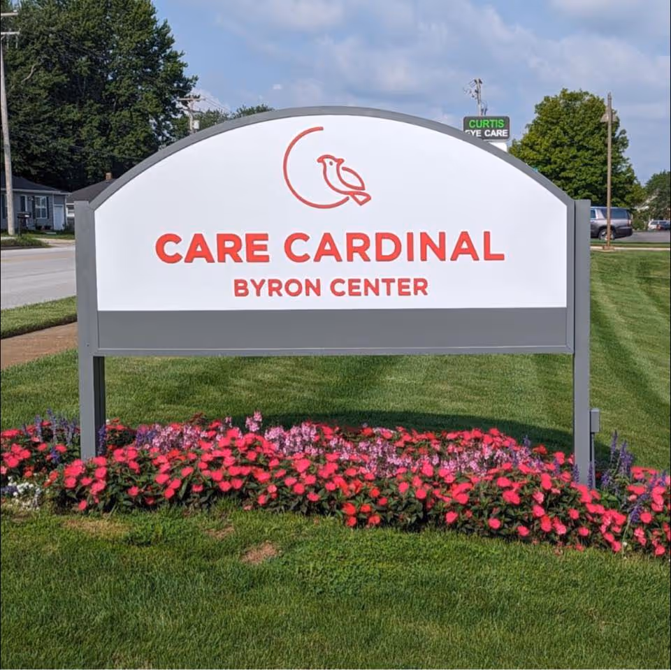 Outdoor sign for Care Cardinal Byron Center with a red cardinal bird logo above the text, surrounded by green grass and pink flowers, with a street and trees in the background under a partly cloudy sky.