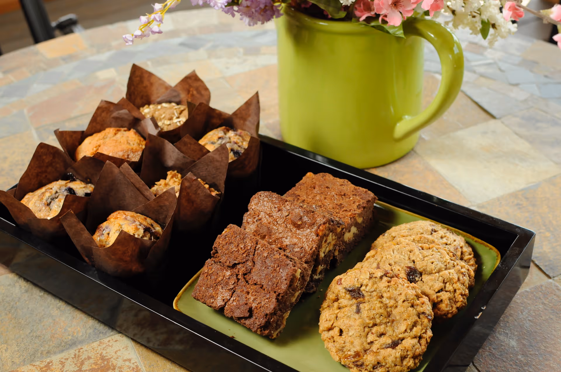 A tray with an assortment of baked goods including muffins in brown paper liners, brownies, and oatmeal cookies with raisins, placed on a tiled table next to a green pitcher holding a bouquet of flowers.