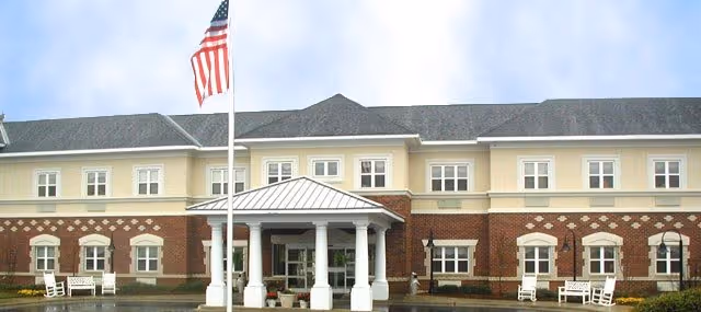 Front entrance of a two-story brick and stucco senior living building with a covered portico, American flag, and rocking chairs on the porch.
