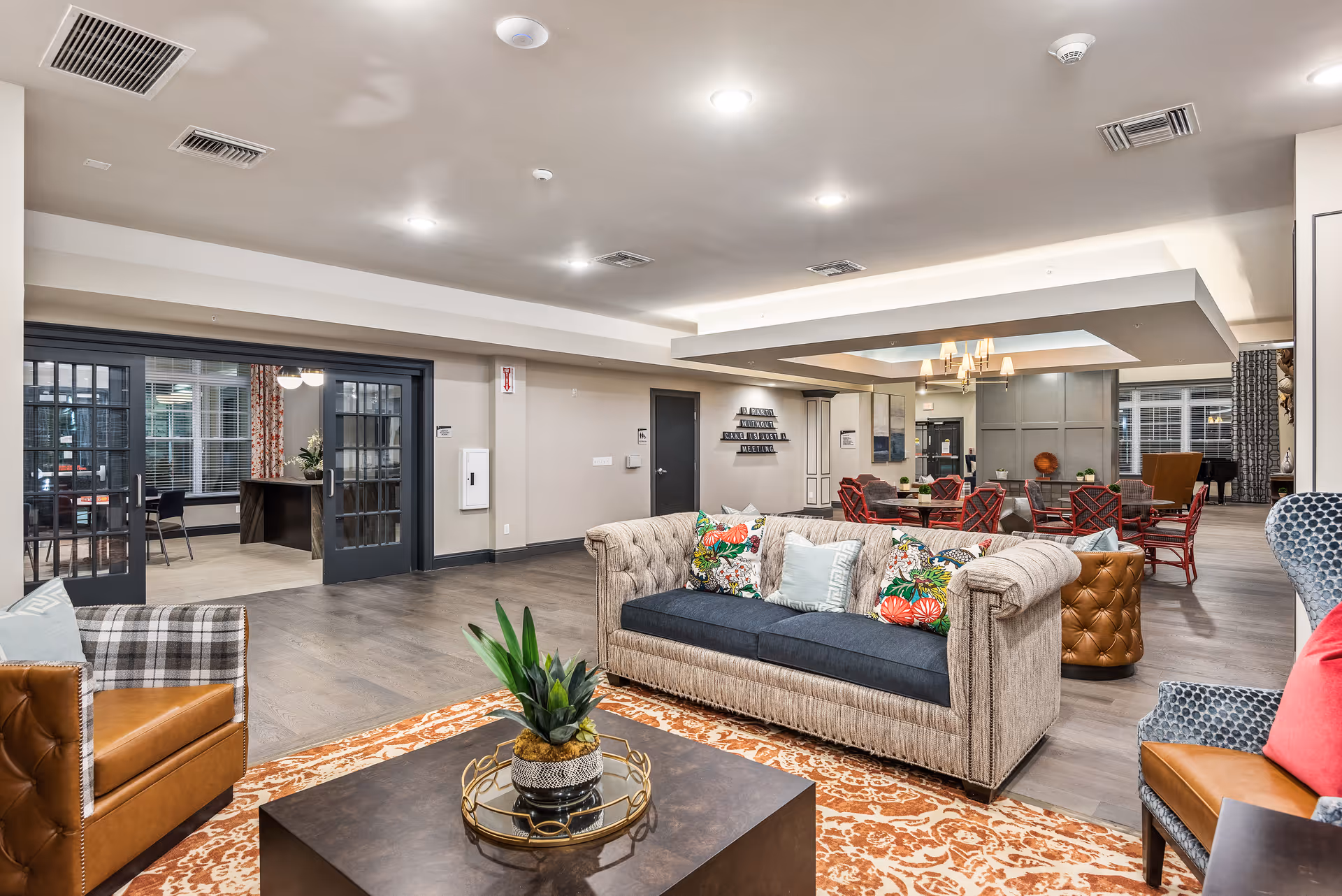 A spacious and well-lit senior living common area featuring a beige sofa with colorful floral pillows, two leather armchairs, a patterned rug, and a dark coffee table with a decorative plant. In the background, there are multiple seating areas with red chairs and tables, a chandelier, and large windows with curtains. The room has light-colored walls and a wooden floor, with a doorway leading to another room with glass double doors.
