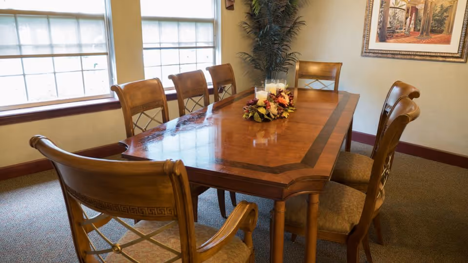 A wooden dining table with six matching chairs in a well-lit room. The table has a decorative centerpiece with candles and flowers. There are two large windows with blinds partially drawn, a potted plant in the corner, and a framed picture on the wall.