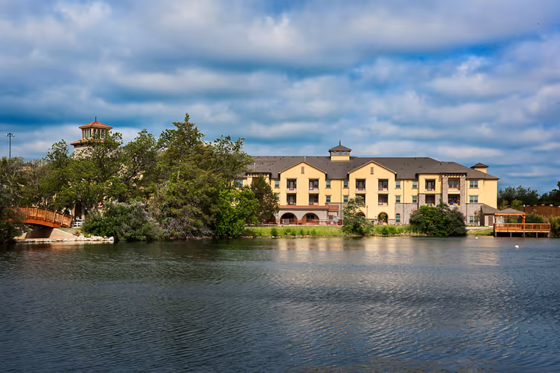 A large multi-story building with a beige and stone exterior situated behind a body of water. The building has multiple balconies and a tower-like structure on the left side. Trees and greenery surround the building, and a wooden bridge is visible on the left side. The sky is partly cloudy.