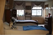 A nursing facility bedroom with two single beds, one in the foreground with a blue blanket and stuffed animals, and another in the background near two windows with curtains. The room has wooden furniture including a dresser, shelves, and a chair, with personal items and decorations on the shelves and windowsills.