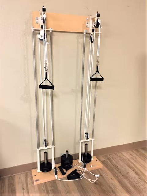 A wall-mounted pulley weight machine with two handles and adjustable weights, placed against a beige wall on a wooden floor.