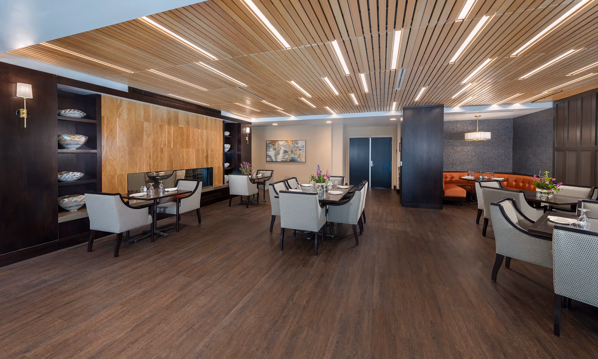 Spacious modern dining room with wood-paneled ceiling and floor, multiple dining tables and upholstered chairs, built-in shelving and a curved booth.
