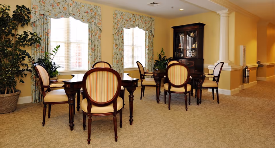 Bright senior living common room with wooden tables and upholstered chairs by windows and a china cabinet.