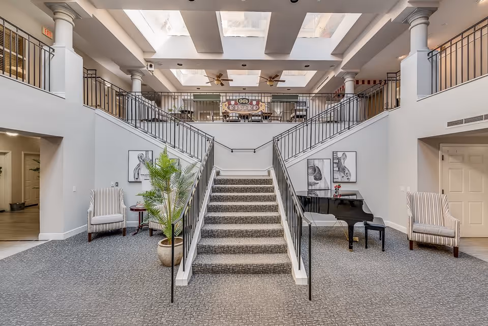Spacious senior living facility interior with a central carpeted staircase leading to an upper level. The area features two striped armchairs on either side of the staircase, a black grand piano with a small bench, potted plants, and framed artwork on the walls. The upper level has a railing and a sign that reads 'GG's Bistro'. The ceiling has multiple skylights allowing natural light to brighten the space.