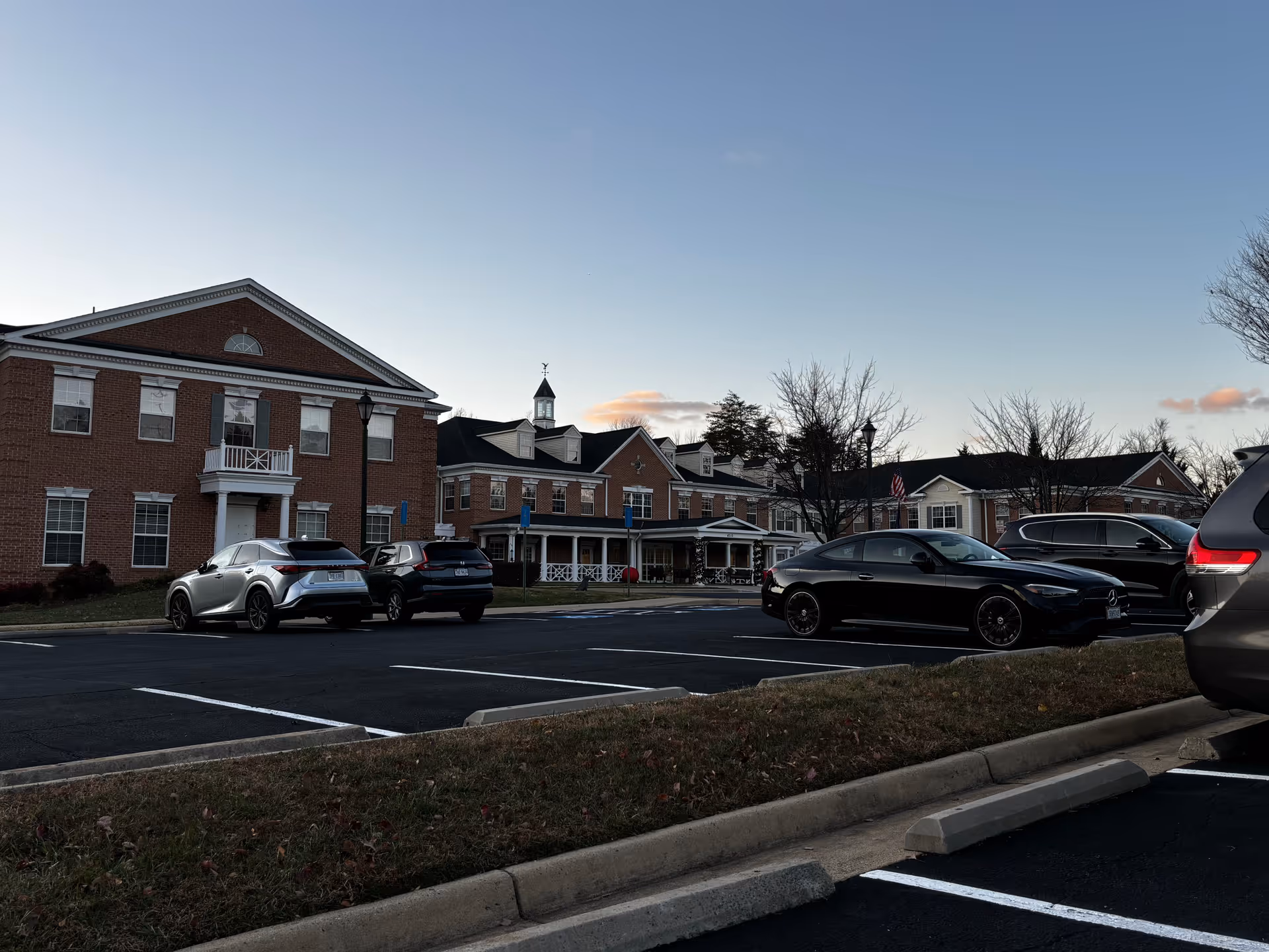 Exterior view of a senior living facility named The Gardens at Fair Oaks during dusk, showing a large brick building with multiple windows, a small cupola on the roof, and a parking lot with several parked cars in the foreground.