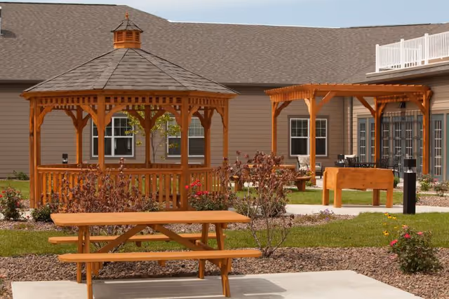 Outdoor area of Lutheran Life Villages - The Village at Inverness featuring a wooden gazebo, a picnic table, a pergola with seating, and landscaped garden beds with shrubs and flowers in front of a beige building with multiple windows.