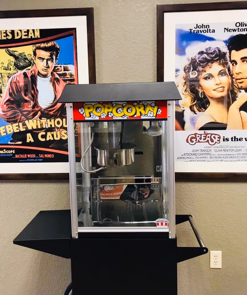 A popcorn machine on a cart stands against a wall between framed movie posters for Rebel Without a Cause and Grease.