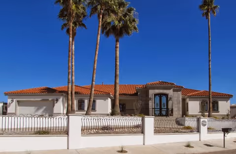 Front exterior view of a single-story building with a red tile roof, beige walls, and a decorative wrought iron gate and fence. There are four tall palm trees in front of the building under a clear blue sky.