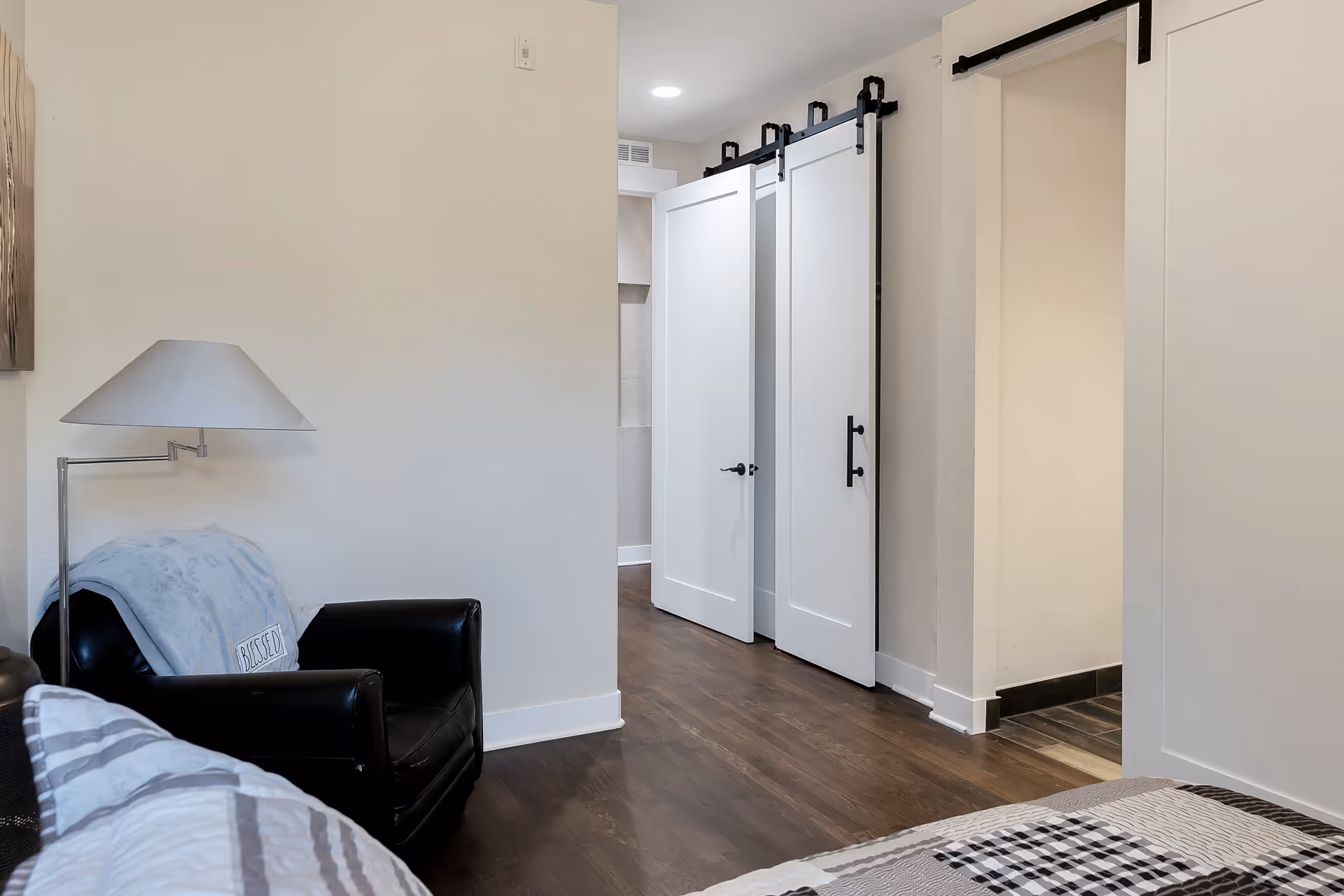 Interior view of a senior living facility bedroom showing a black armchair with a light blue blanket and a pillow, a modern floor lamp, a bed with a patterned quilt, dark wood flooring, and white sliding barn doors leading to another room.