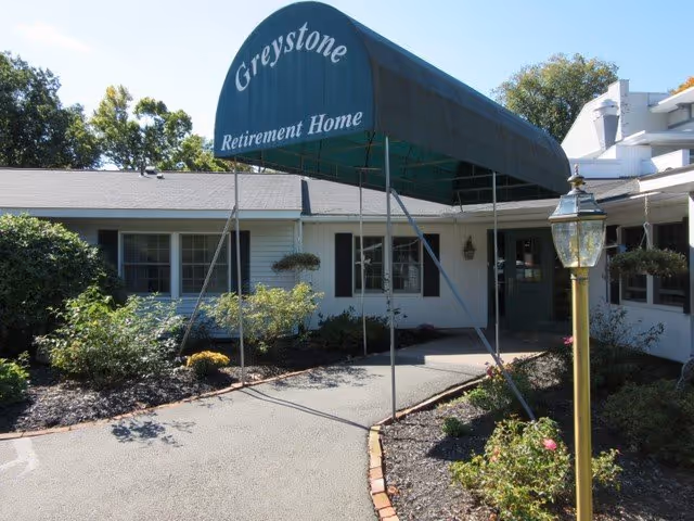 Front entrance of Greystone Retirement Home with a green canopy over a paved driveway and surrounding landscaping.