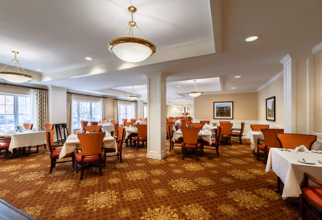 A spacious dining room with multiple tables covered in white tablecloths and set with napkins and silverware. The room features orange upholstered chairs, patterned carpet, large windows with curtains, and ceiling light fixtures.