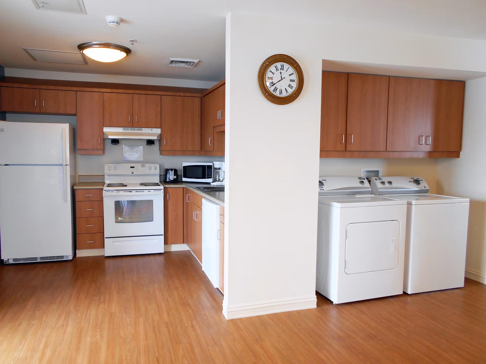 Interior view of a kitchen and laundry area in a senior living facility. The kitchen features wooden cabinets, a white refrigerator, white stove with an oven, microwave, toaster, and coffee maker. Adjacent to the kitchen is a laundry area with a white washing machine and dryer, also with wooden cabinets above. A round wall clock is mounted on a white wall separating the two areas. The floor is a light wood laminate.