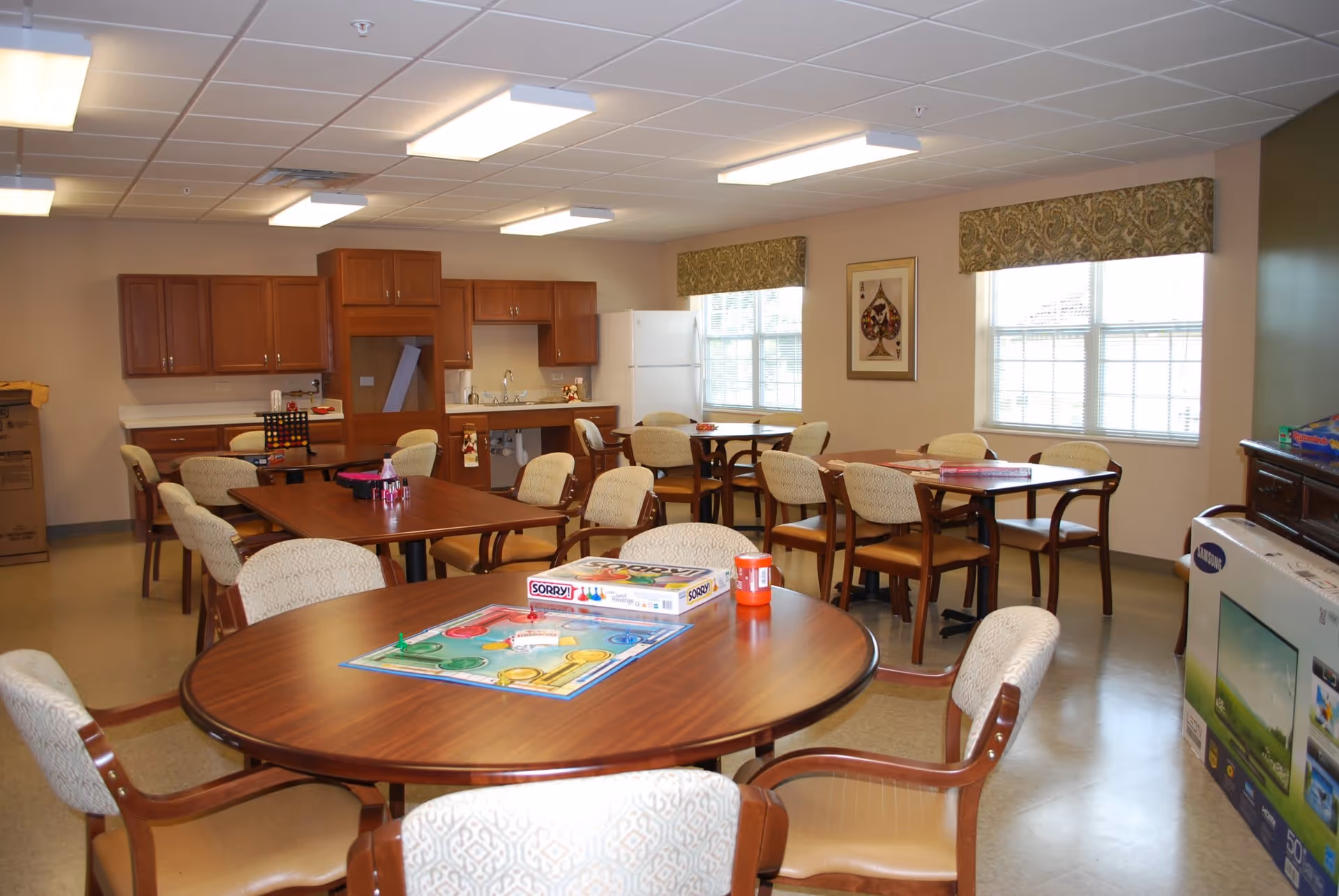 Communal dining/activity room with multiple round and rectangular tables and chairs, a kitchenette in the back, and board games on the tables.
