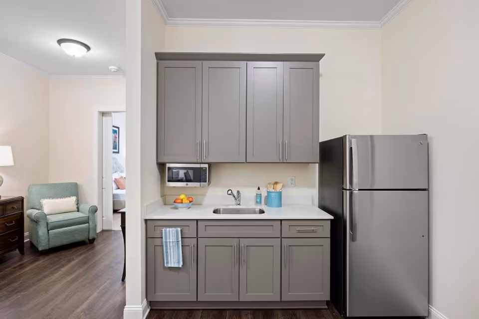 A small kitchen area with gray cabinets, a stainless steel refrigerator, a microwave mounted under the upper cabinets, a sink with a soap dispenser and utensils on the counter, and a blue towel hanging on a cabinet handle. To the left, there is a partial view of a living area with a green armchair, a wooden side table with a lamp, and a doorway leading to a bedroom.