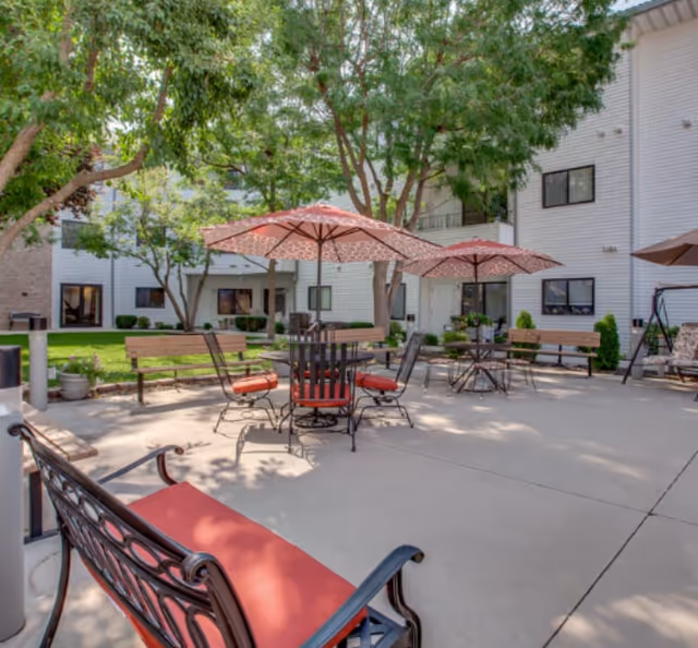 Outdoor courtyard at a senior living facility with patio tables, umbrellas, benches, and surrounding building façades.