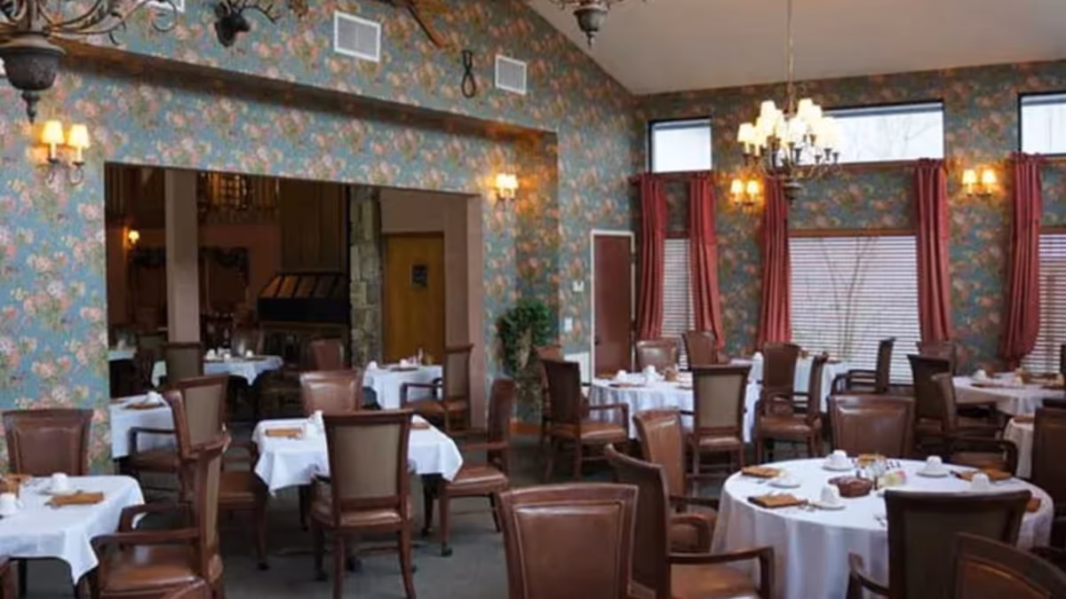 Dining room with multiple round and square tables covered with white tablecloths, set with cups and napkins. The room has floral wallpaper, large windows with red curtains, and several chandeliers and wall sconces providing warm lighting. Brown leather chairs surround the tables.