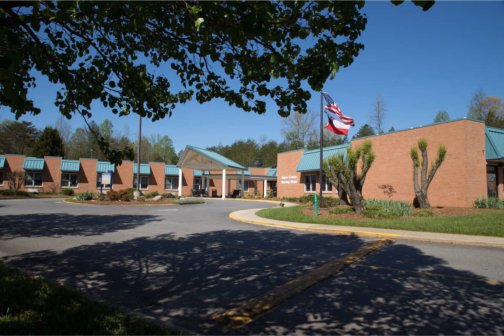 Brick single-story nursing home building with a covered entrance, American flag poles, and a circular driveway under a clear blue sky.