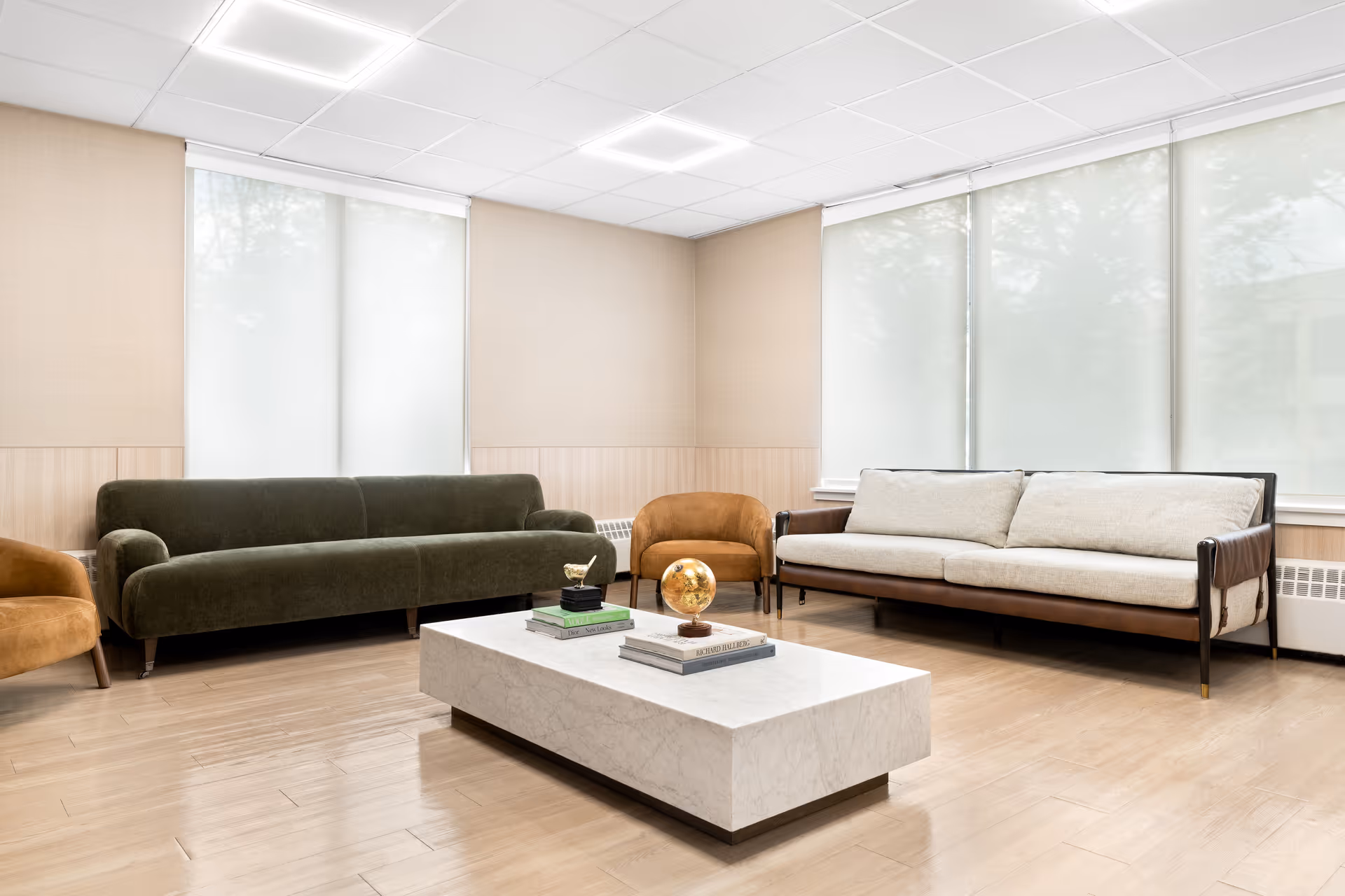 A bright and modern living room with two sofas, one dark green and one light beige with wooden arms, two brown armchairs, and a rectangular white marble coffee table with books and a small globe on it. Large windows with white blinds cover the walls, and the floor is light wood.