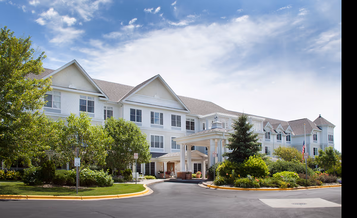 Exterior view of a large, white senior living facility building with multiple windows, a covered entrance, and landscaped greenery including trees and bushes under a partly cloudy sky.