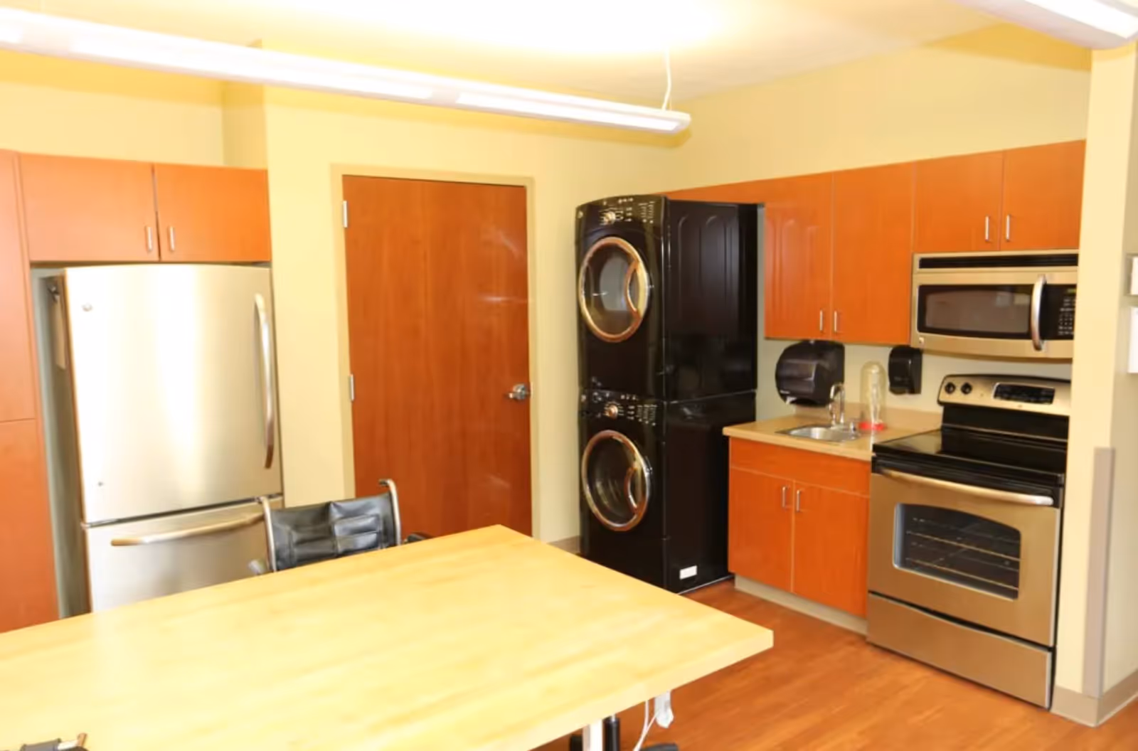 Interior view of a kitchen area with wooden cabinets, a stainless steel refrigerator, stove, microwave, and a stacked washer and dryer unit. There is a wooden table with a black chair in the foreground and a small sink with a soap dispenser on the countertop.