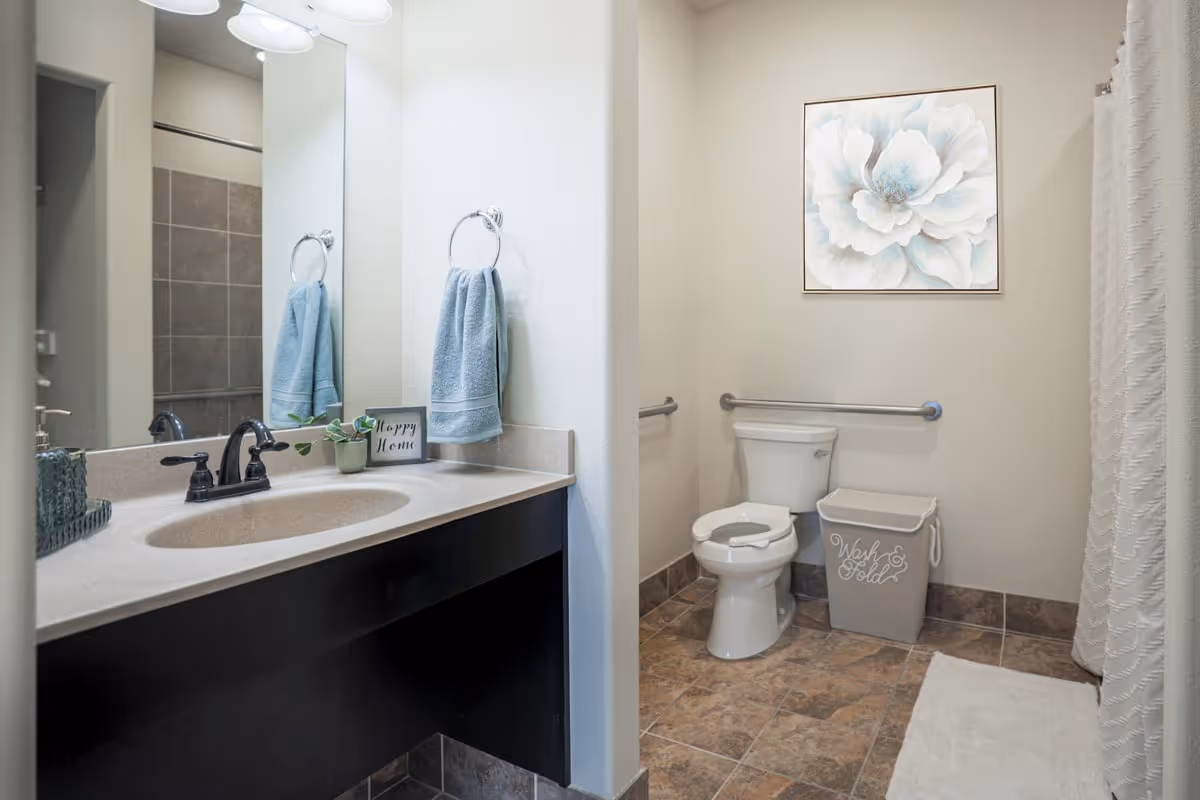 A clean and accessible bathroom with a white toilet, a beige countertop with a sink, a large mirror, a blue hand towel hanging on a ring, a small plant, and a decorative sign that says 'Happy Home'. There is a laundry basket labeled 'Wash & Fold', a white bath mat on the tiled floor, a grab bar next to the toilet, and a large floral painting on the wall.