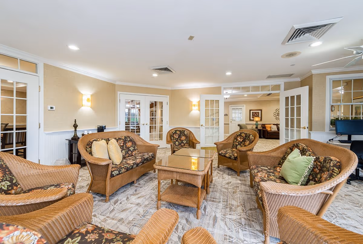 A bright and spacious living room area with wicker furniture including sofas and chairs with floral cushions arranged around a glass-top coffee table. The room has beige walls, carpeted floors, and multiple glass-paneled doors leading to other rooms. There are wall sconces providing warm lighting and a computer workstation visible in the background.