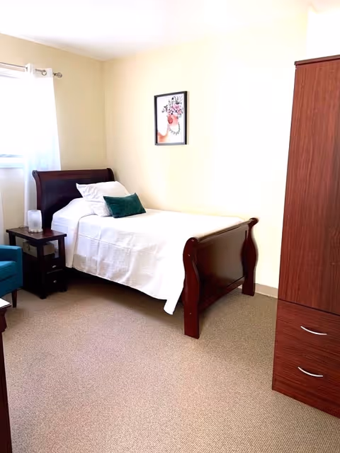 A neatly arranged bedroom with a single wooden bed covered in white bedding and a green pillow. There is a small wooden nightstand with a lamp next to the bed, a blue armchair partially visible, a wooden wardrobe with drawers, and a framed floral artwork hanging on the light-colored wall. A window with white curtains allows natural light into the room.