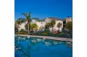 Low-rise apartment building with palm trees and a swimming pool reflecting the building under a clear blue sky.