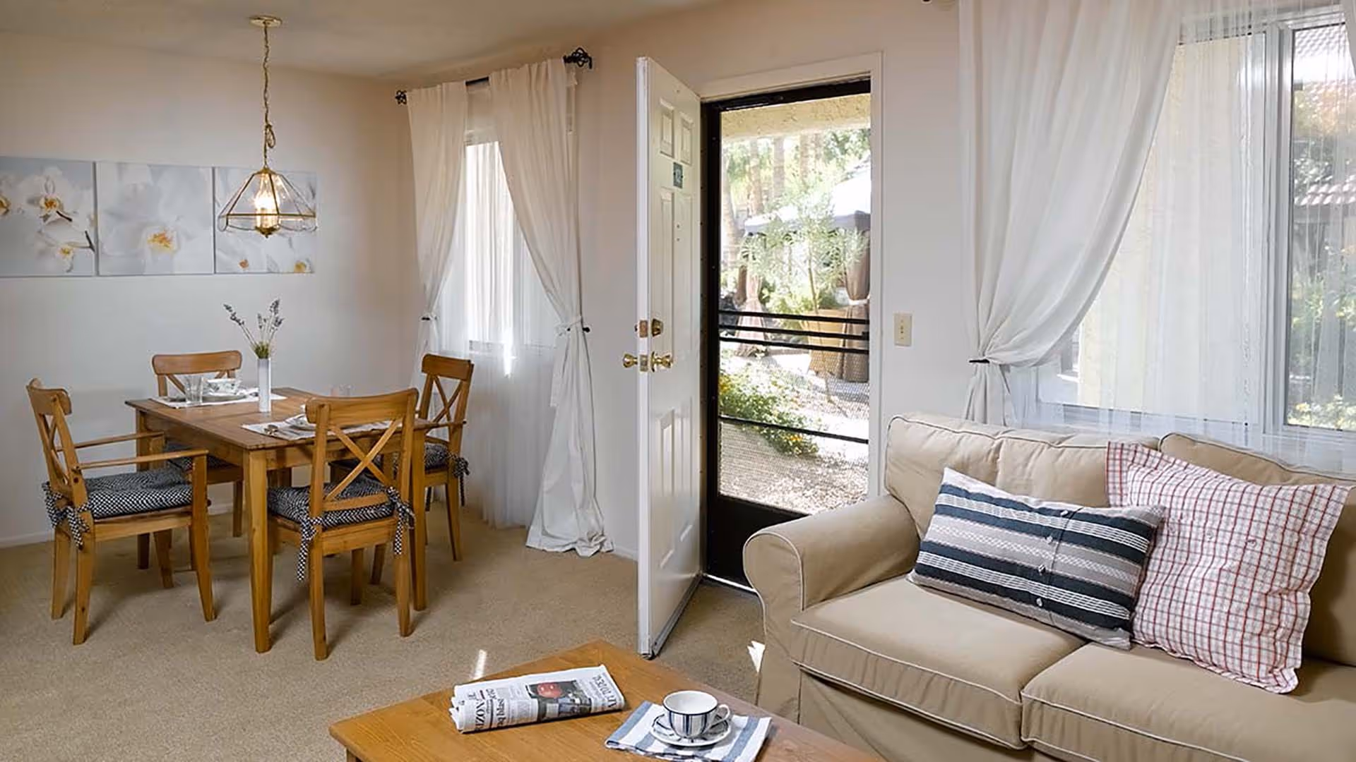 Sunny open living and dining area with a beige sofa, wooden dining table, and a door opening to an outdoor patio.