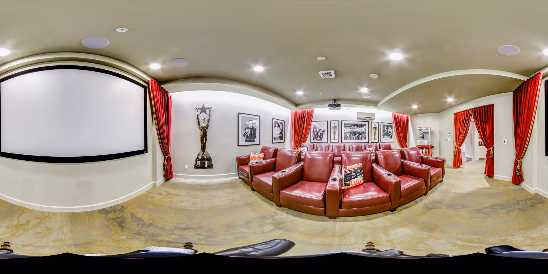 A cozy home theater room with red leather reclining chairs arranged in rows facing a large white projection screen. The room features red curtains on the walls, framed black and white photos, a large trophy statue, and a popcorn machine in the back corner. The ceiling has recessed lighting and a projector mounted in the center.